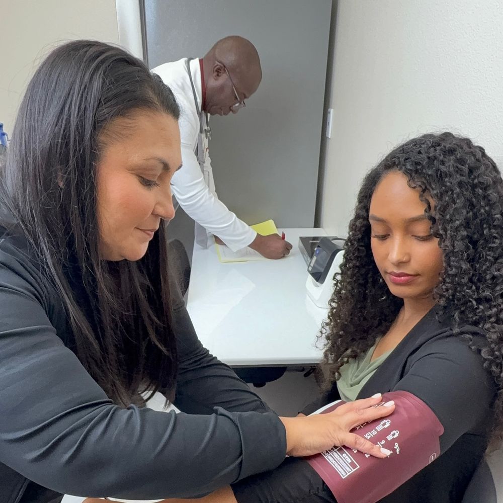Medical assistant placing a blood pressure cuff on a patient at Upper Valley Wellness Clinic while the doctor works at the desk behind them