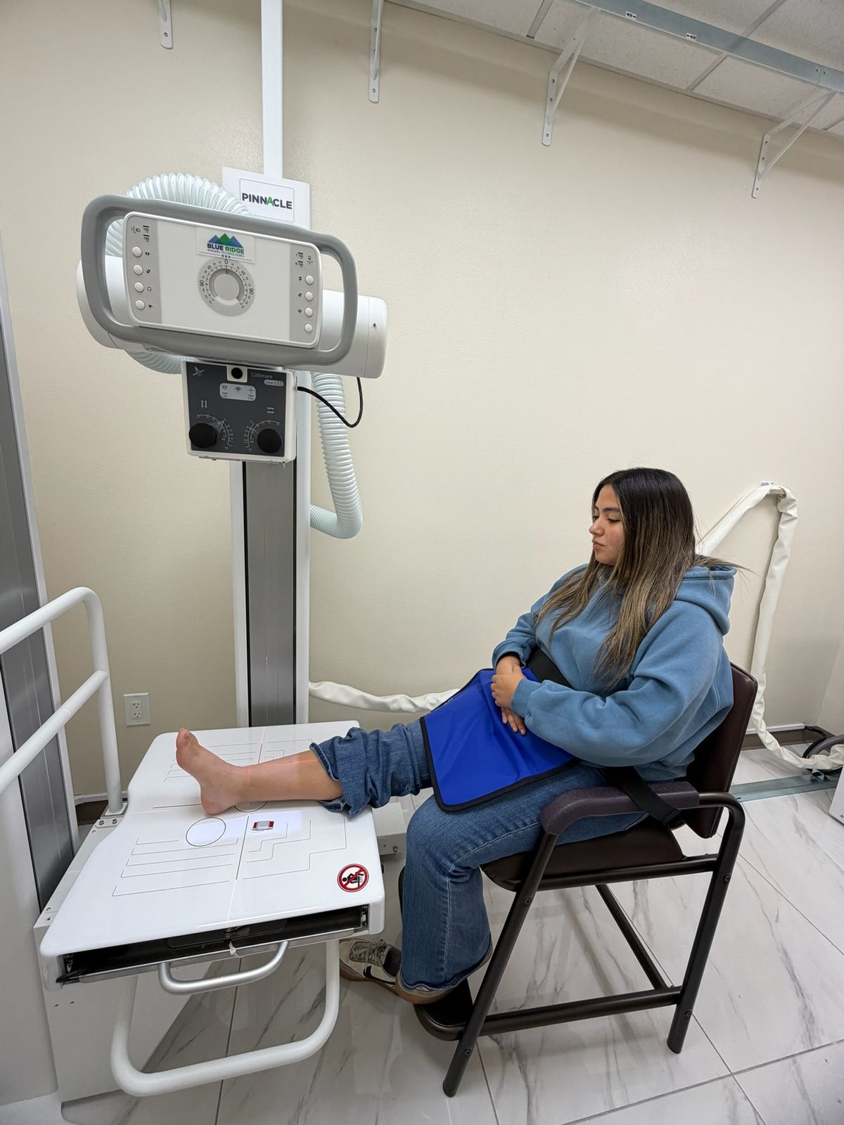 Patient seated with her foot on the X-ray table, wearing a protective apron, in the Upper Valley Urgent Care X-ray room